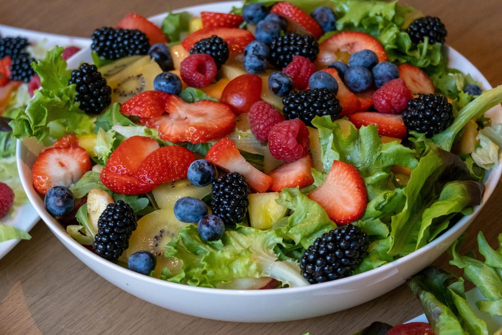 A white bowl filled with lettuce, strawberries, raspberries, blueberries, blackberries, kiwi, and pineapple