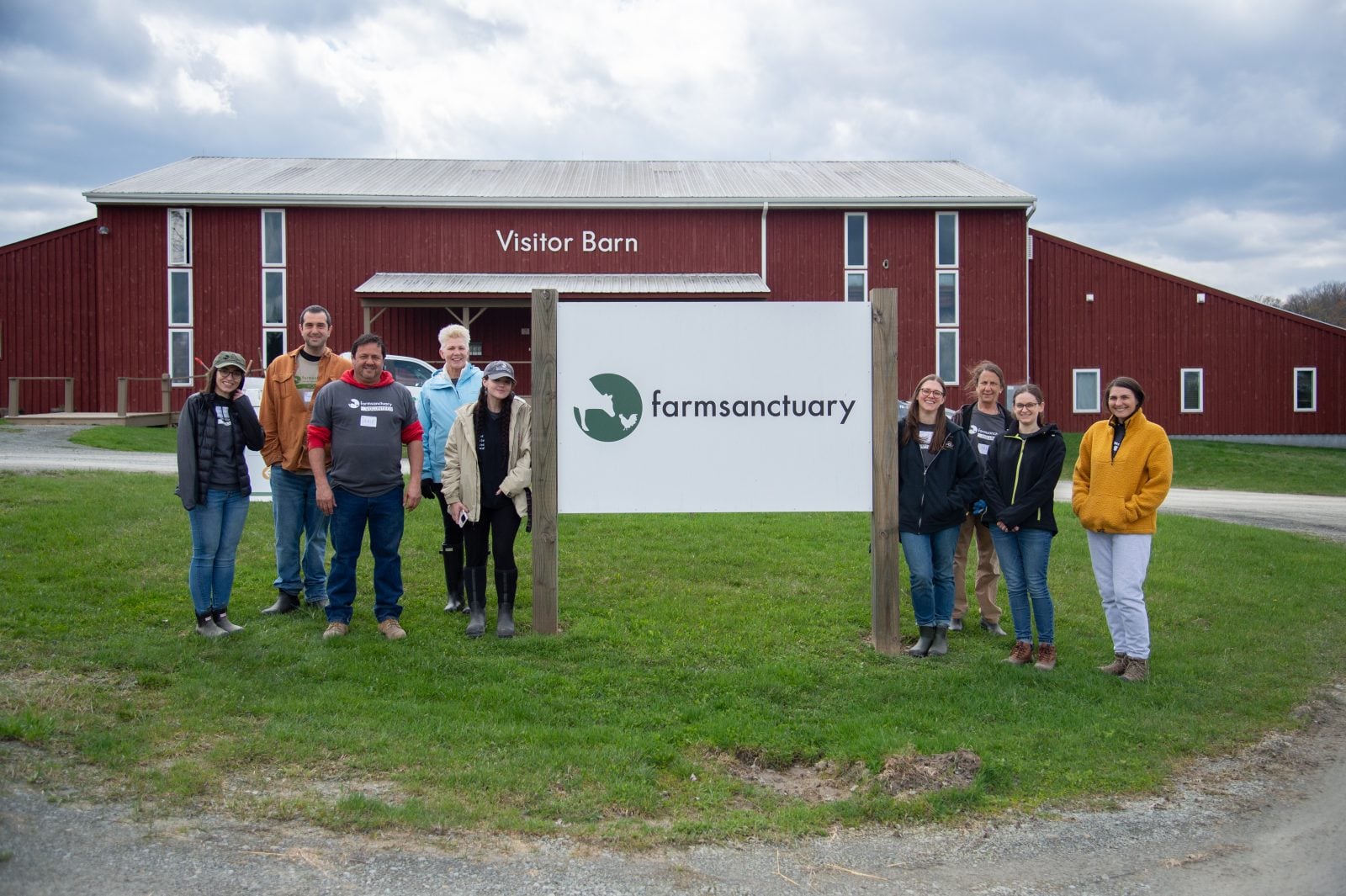 Nine volunteers stand on either side of sign reading "Farm Sanctuary," in front of red barn reading "Visitor Barn"