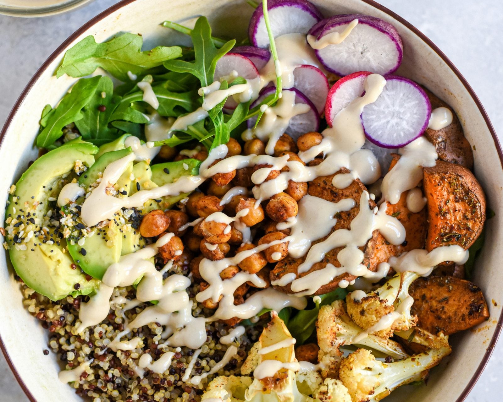 Bowl of greens, avocado, rice, cauliflower, sweet potato, radish, seeds, and a drizzle of sauce