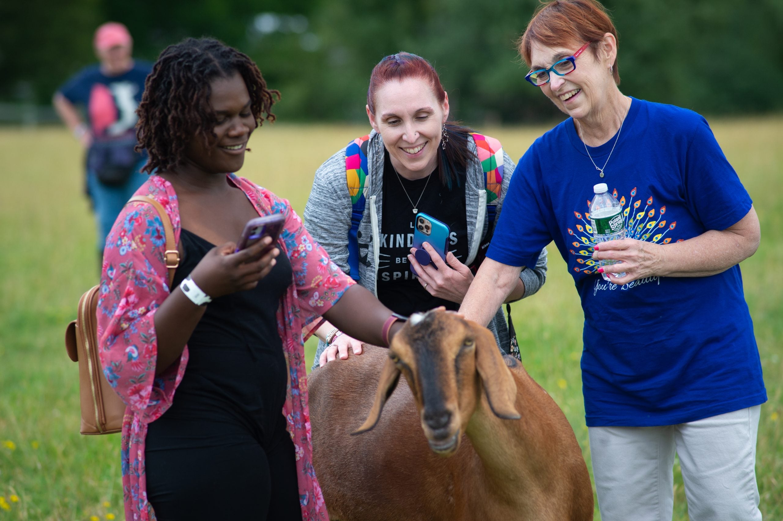 Three smiling guests pet rescued goat Halbert in pasture at Farm Sanctuary