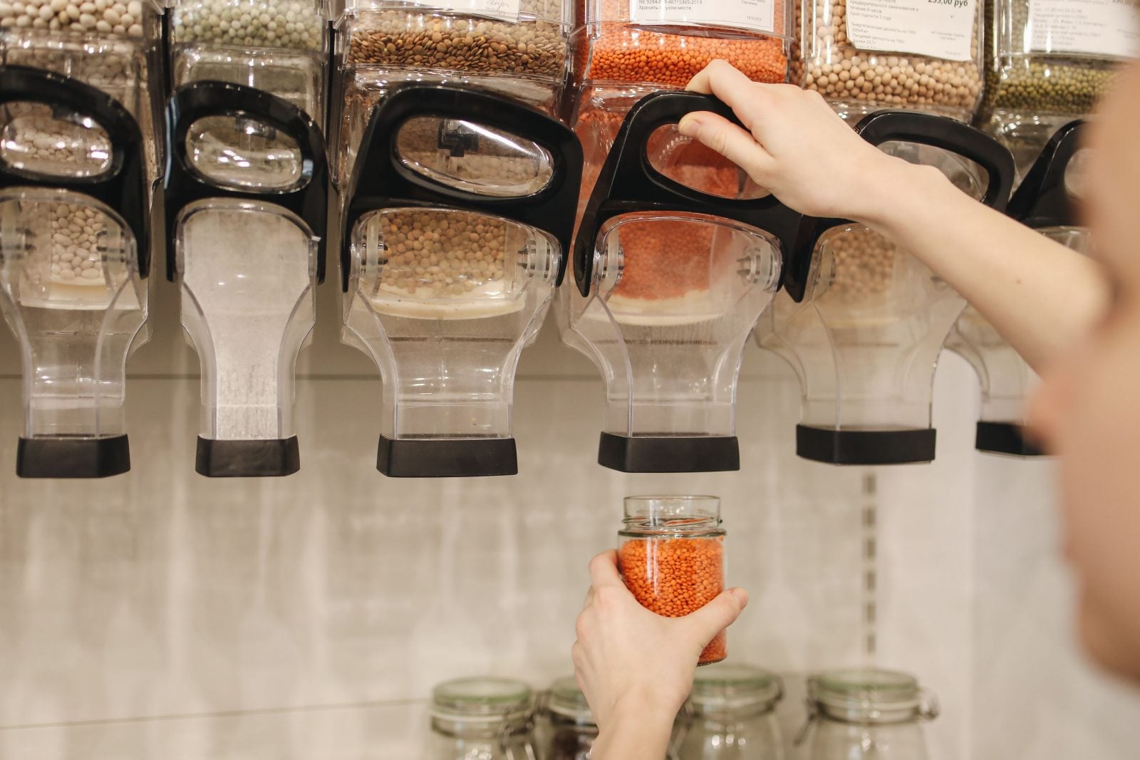 A person reaches up to pour orange lentils from a bulk dispenser into a glass jar