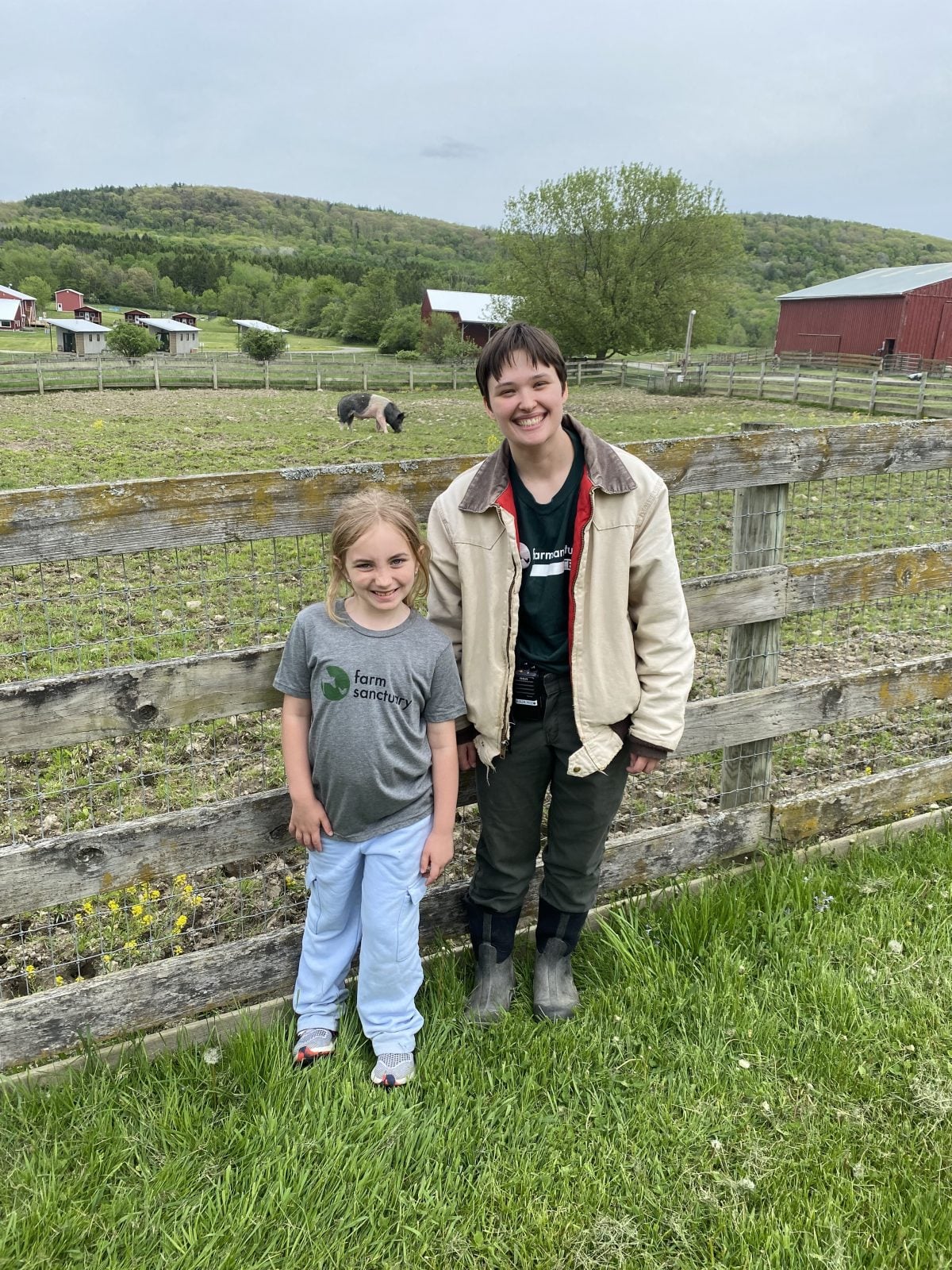 Young student smiles with Farm Sanctuary staff Julia Marotto in front of fence, pasture, and barns