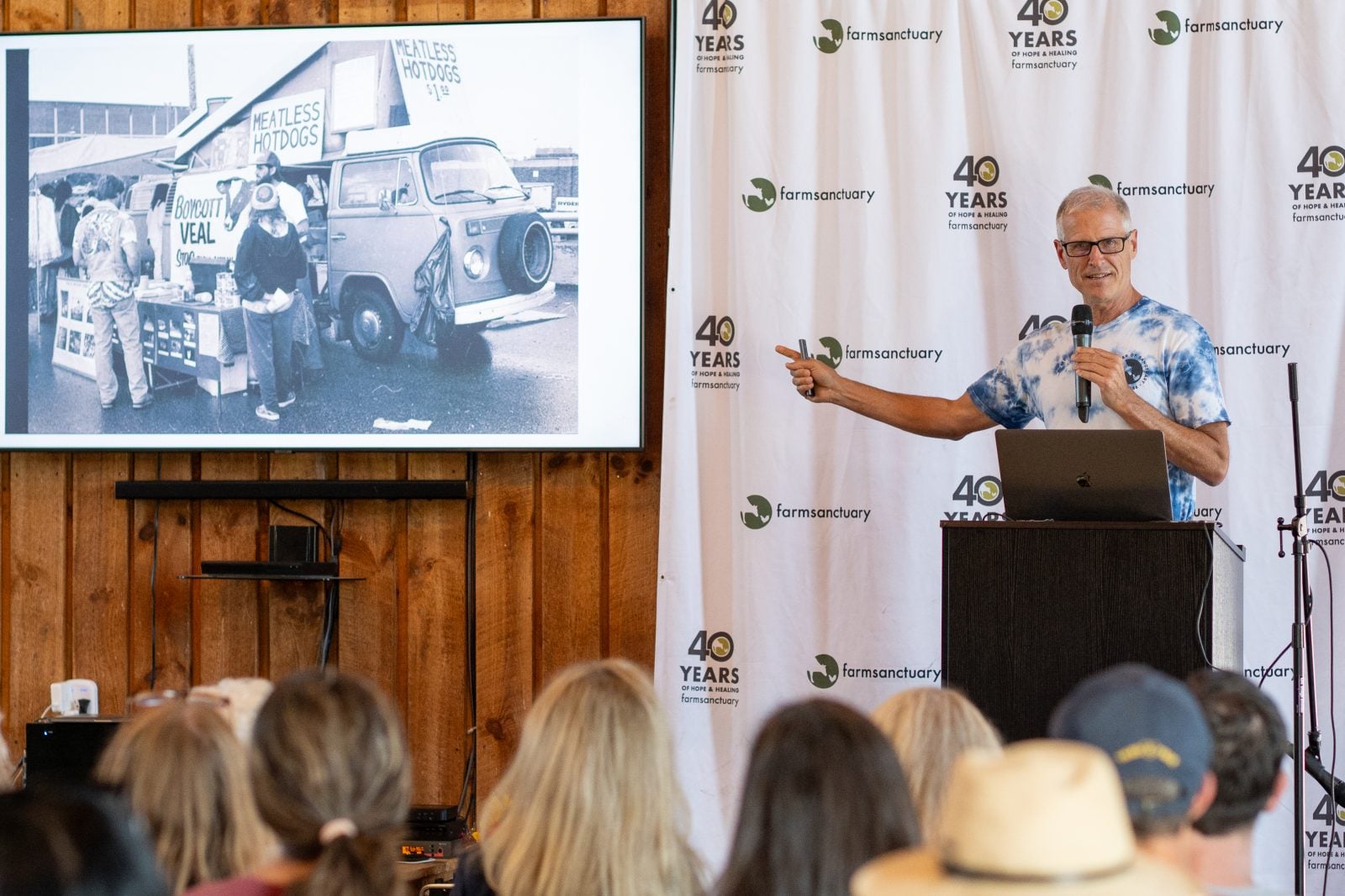 Gene Baur gestures to black-and-white photo of activists outside a VW van with signs reading "meatless hot dogs" and "boycott veal"