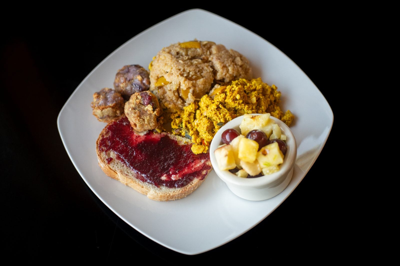 A white plate on black background with tofu scramble, fruit cup, toast with jelly spread, biscuit